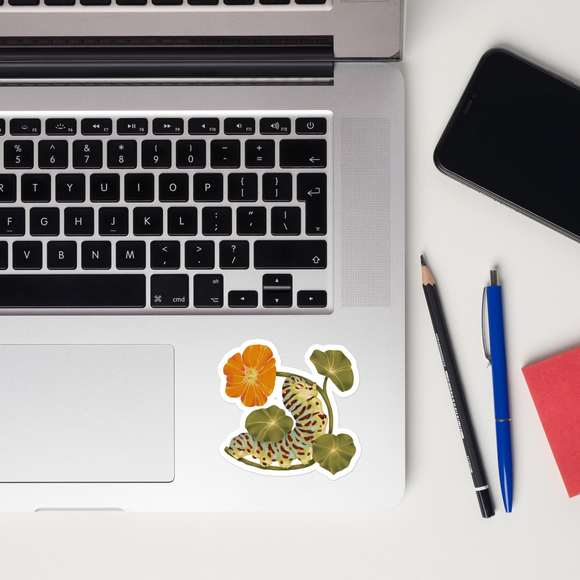 Laptop with a floral sticker on a white surface, next to a phone, pencil, and pen.